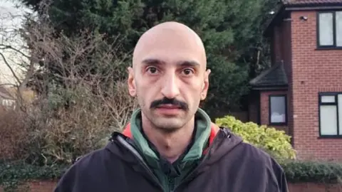 handout Farid Vahidi poses on a street with trees behind. He wears as dark coat over a top in the green and red colours of the Iranian flag