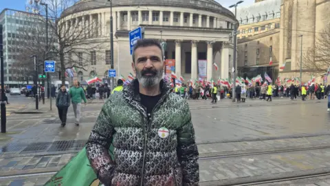 Dainel poses in front of the rally outside the circular Manchester Central Library. He wears a zipped up coat with an Iranina badge and flag flying from his pocket.