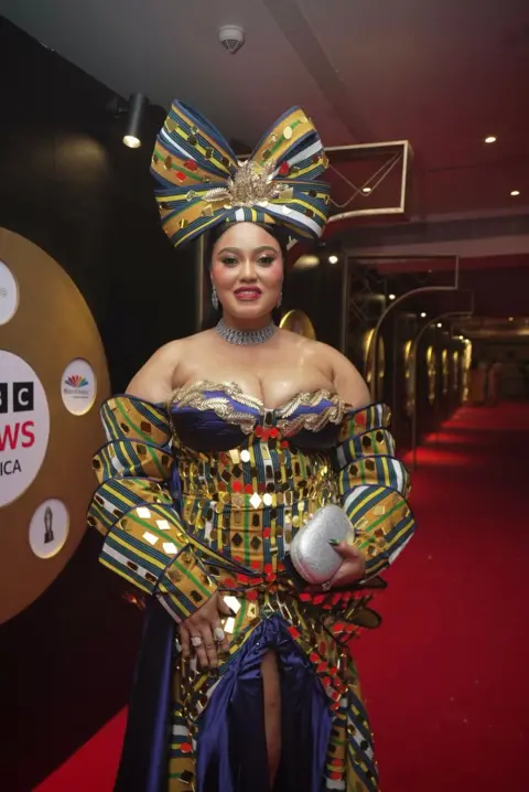 BBC/GIFT UFUOMA Reality TV star OJ Posharella graces the red carpet wearing a multi-layered, colorful costume of her own design and a giant bow on her head.