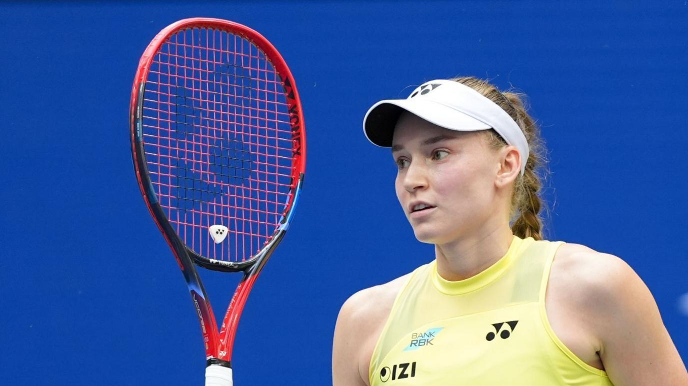 Elena Rybakina, in a yellow outfit, white visor and holding a largely red and black racquet
