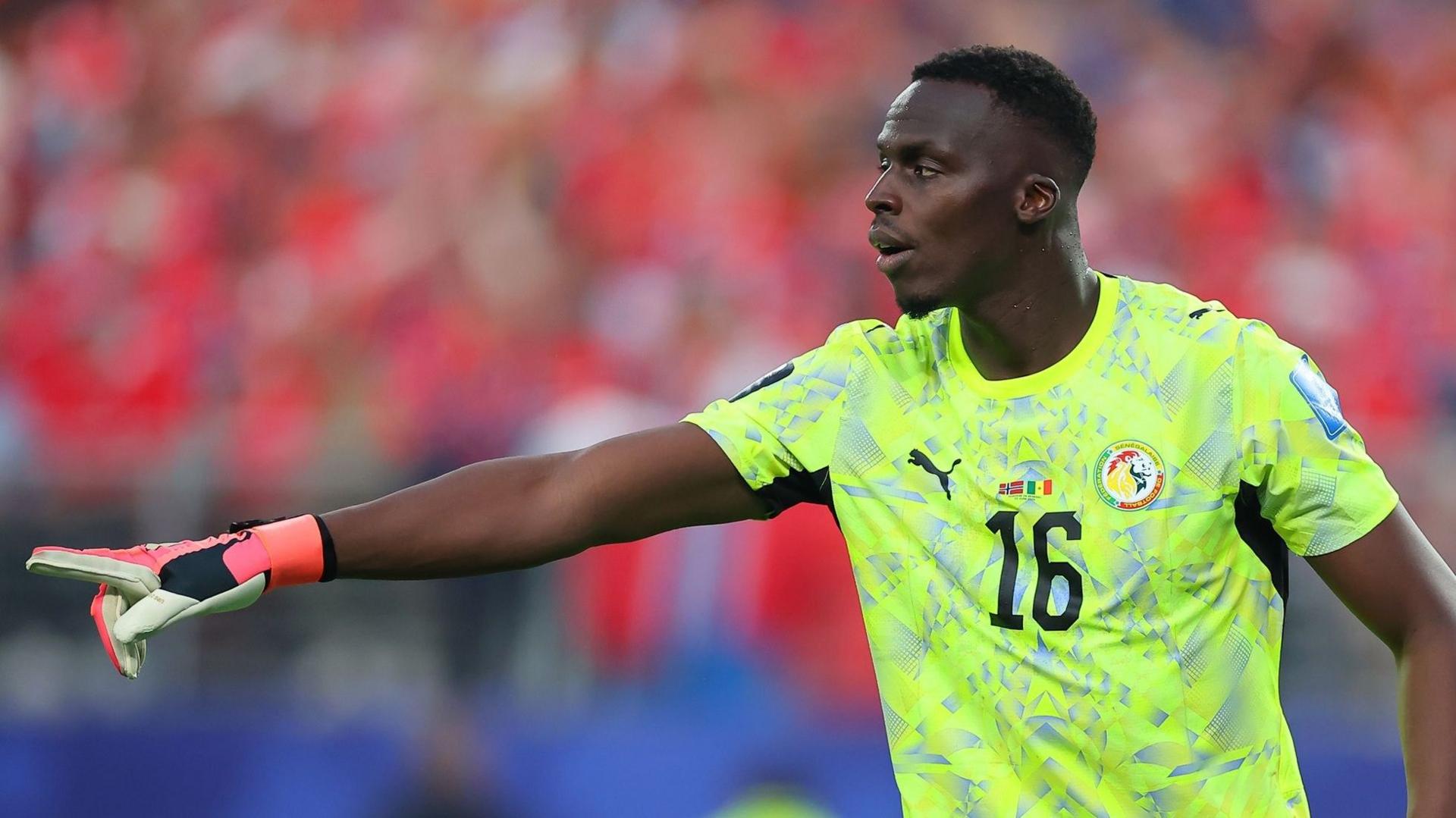 Edouard Mendy, visible from waist up wearing a pink and black goalkeeping glove on his right hand and a luminous yellow Senegal shirt with a black number 16 on the chest, points his right arm out in front of him at chest level during a football match. A full stand of supporters can be seen out of focus in the background