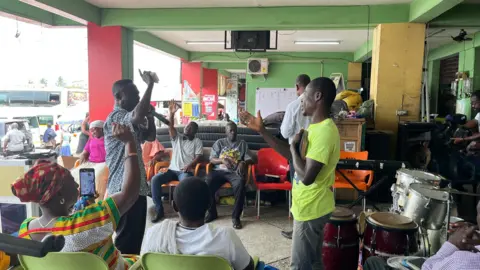A group of people in a large room, which is open on one side, where buses can be seen. They are gesturing with their hands, while preaching. A drum kit is seen behind them.