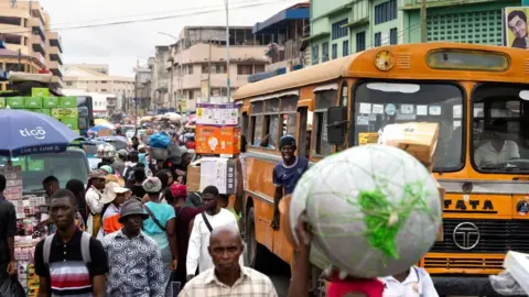 Getty Images A crowded street showing people walking near a yellow Tata commercial bus. In the foreground, an older man is walking towards another man who is carrying a large, white-strapped bundle on top of their head.